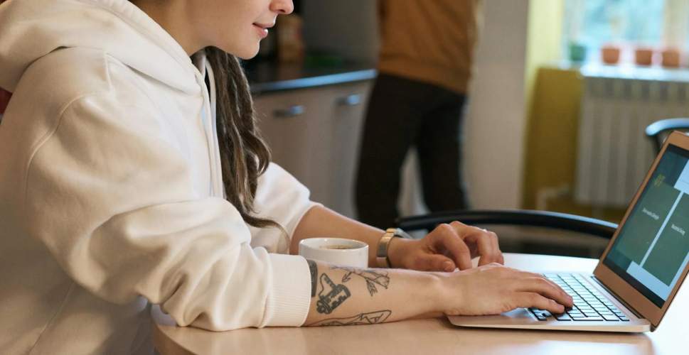 遠端工作情境照片，年輕女性穿著白色帽T在家中使用筆記型電腦工作，桌上放著咖啡杯